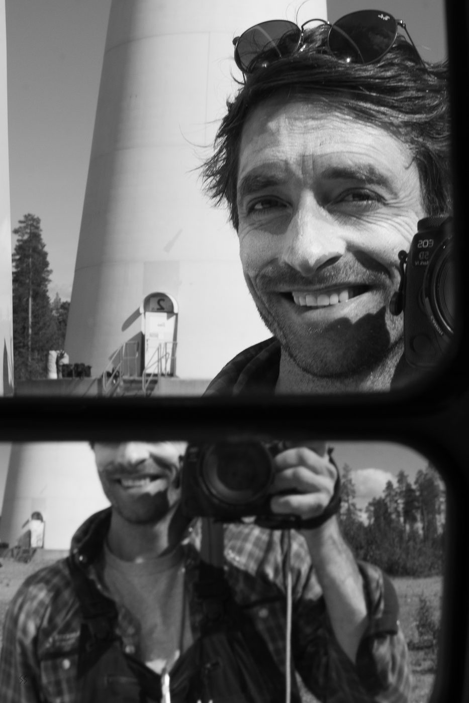 Simon Downing reflected in a vehicle mirror at a wind farm, camera in hand, a turbine tower behind