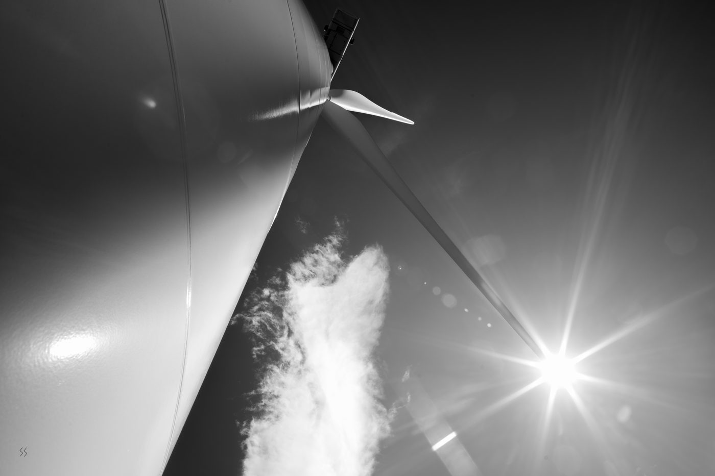 A wind turbine nacelle and blade seen from below against a sunburst