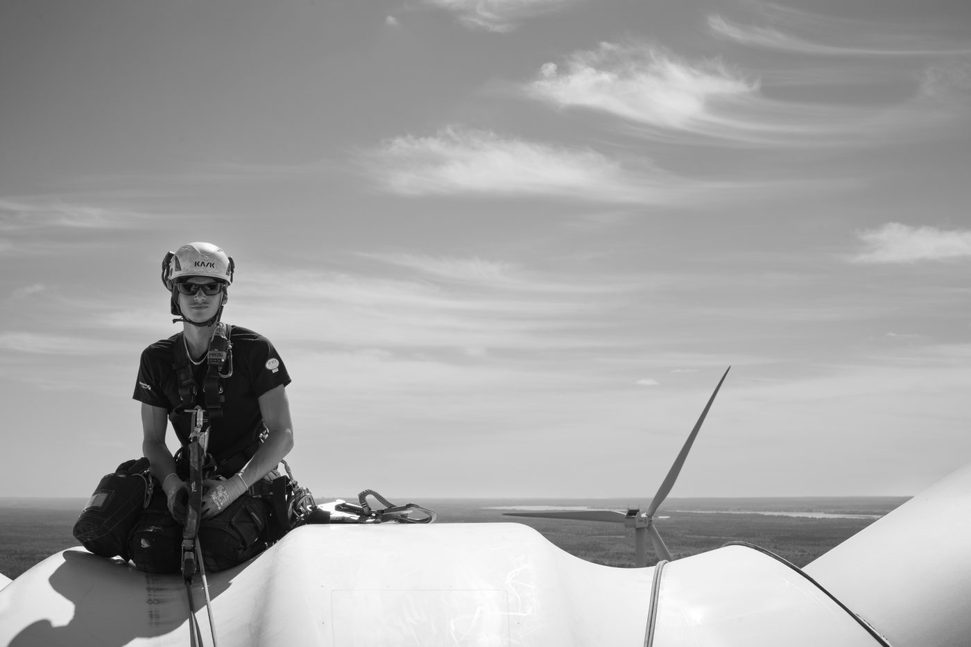 Rope access technician in helmet and harness sitting on the nacelle of a wind turbine, a second turbine and forest far below